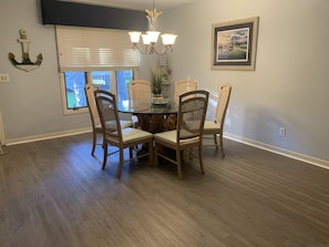 A dining area with a round table, four wicker chairs, a chandelier, and a window with blinds.