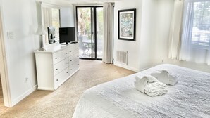 Quarto com cama, penteadeira, televisão e porta de vidro corrediça para uma varanda.
