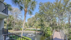 Um deck de madeira com guarda-corpo, com vista para um lago com uma fonte, cercado por vegetação exuberante e palmeiras.