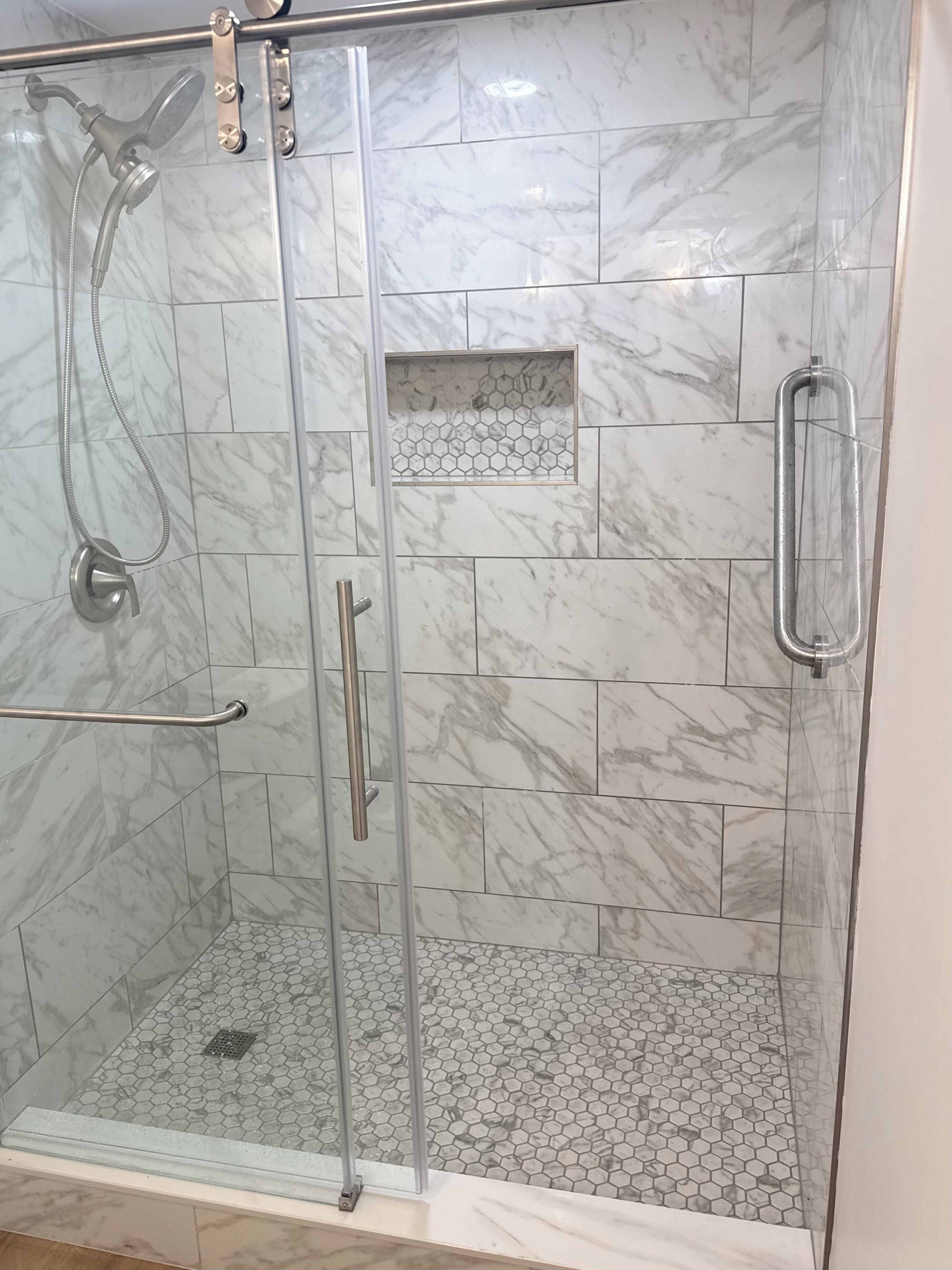 Ducha con cerramiento de vidrio, pared de azulejos de mármol y suelo de baldosas hexagonales, con barandilla de metal.