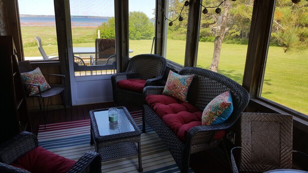 A cozy room with wicker furniture, a striped rug, and a view of a grassy field and water body. - Cozy, private ocean/ beach front cottage with spectacular views in Fortune PEI (Souris)