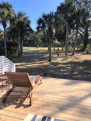 A wooden deck with two lounge chairs and a small table, surrounded by palm trees and a grassy area.