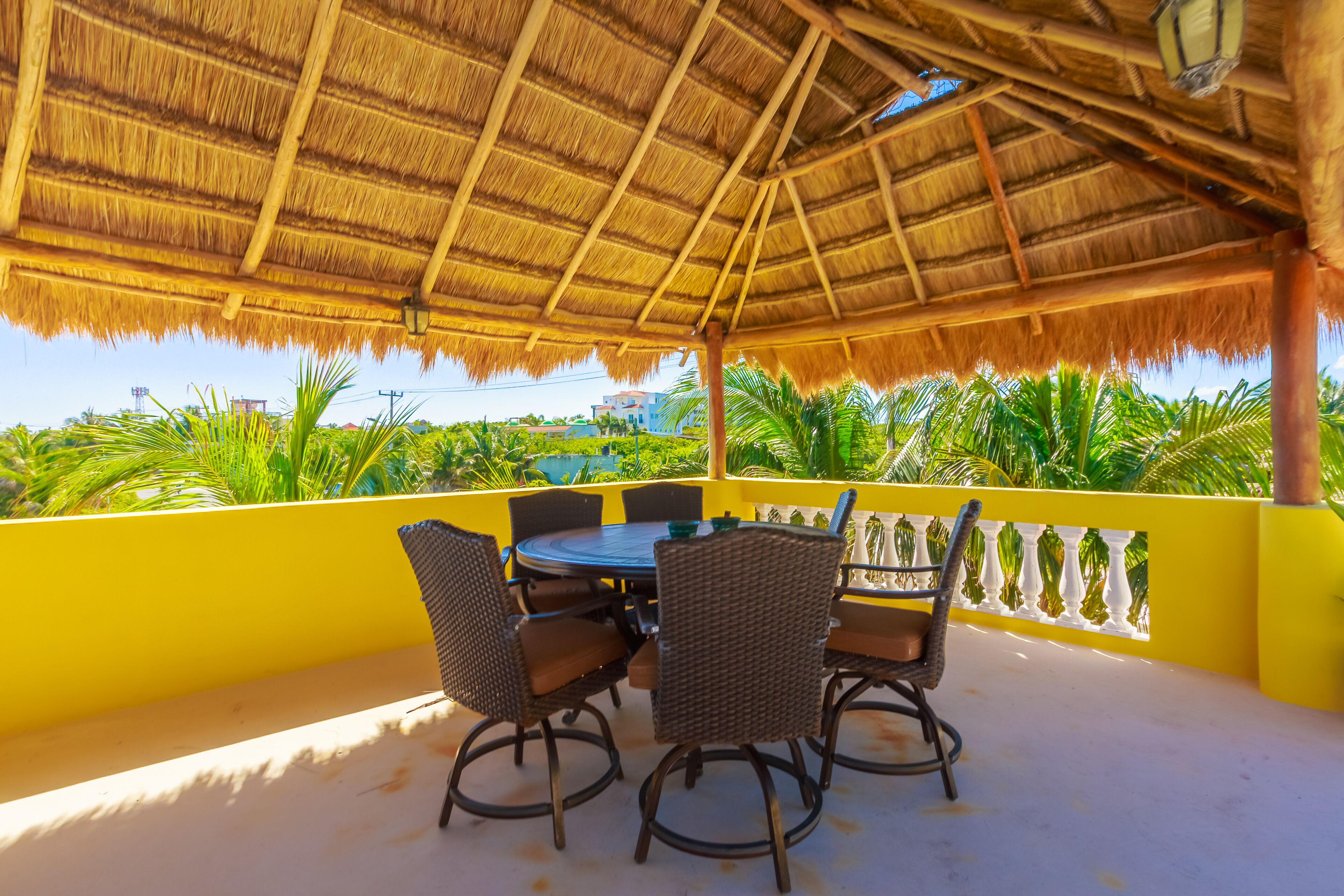 A covered outdoor seating area with a thatched roof, wicker chairs, and a yellow railing.