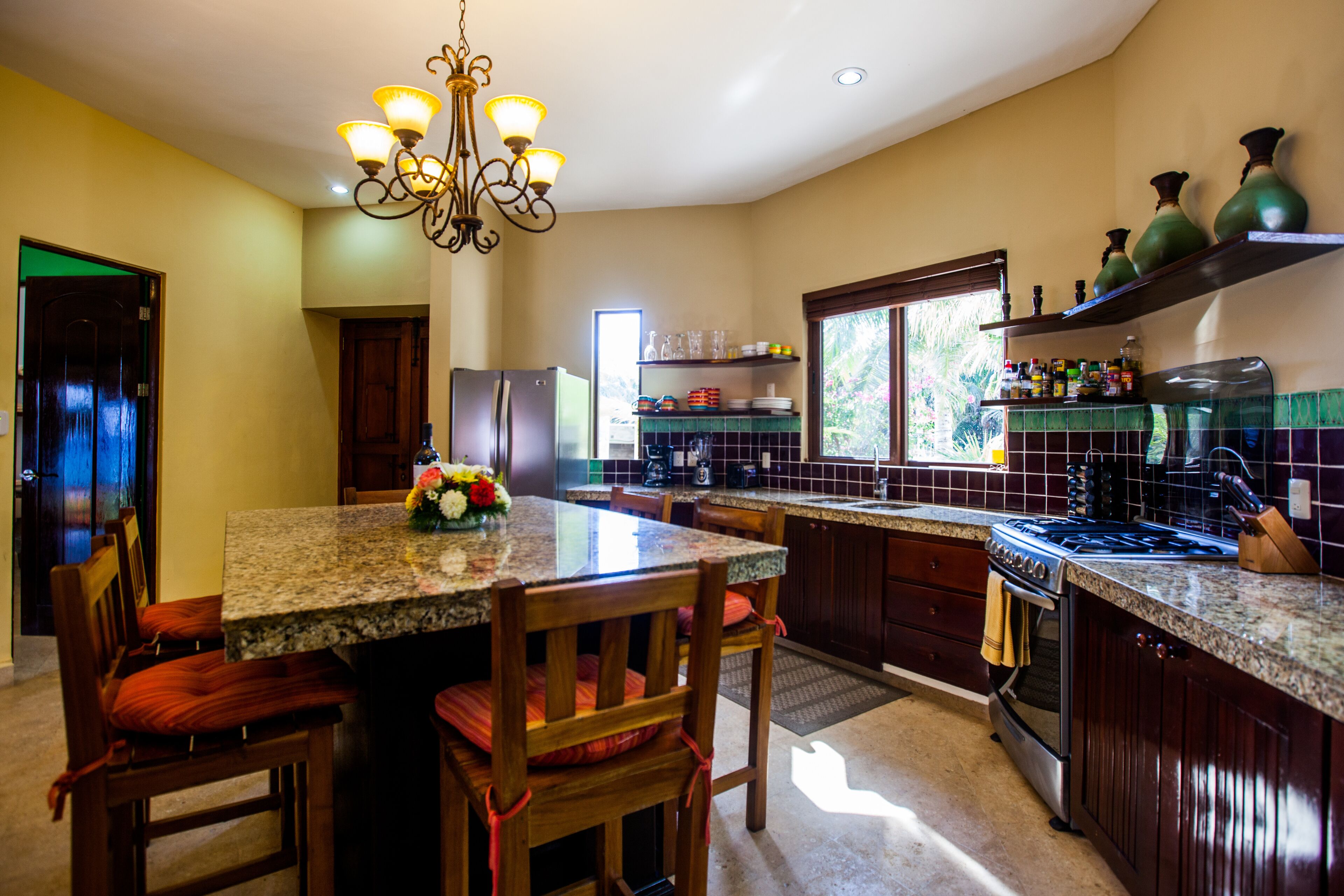 A kitchen with granite countertops, wooden chairs, a chandelier, a refrigerator, and a stove.