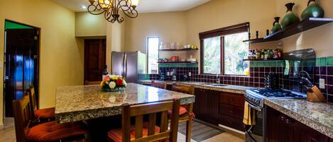 A kitchen with granite countertops, wooden chairs, a chandelier, a refrigerator, and a stove.