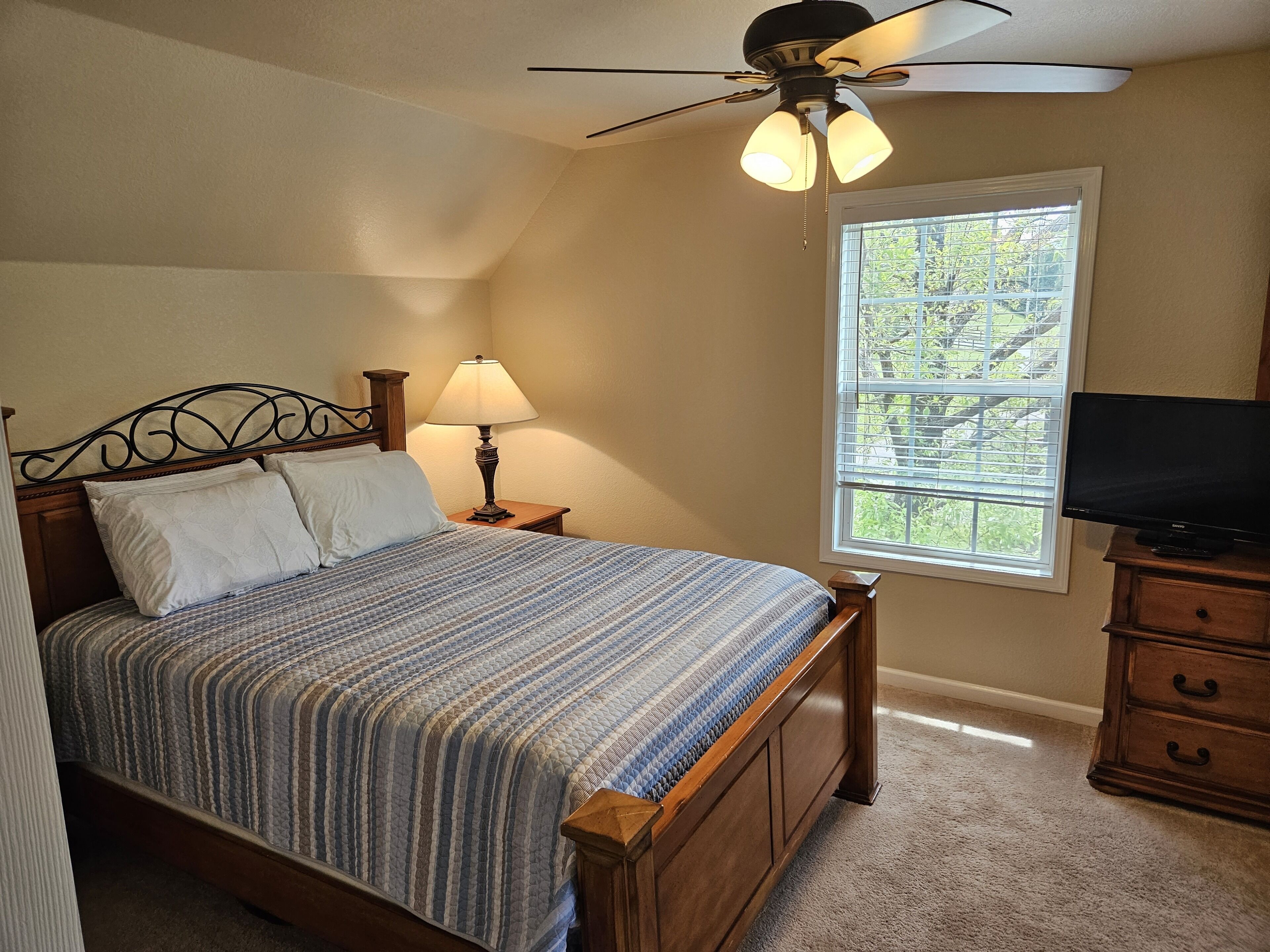 A bedroom with a bed, a nightstand with a lamp, a ceiling fan, a window with blinds, and a TV on a stand.