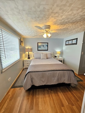 Une chambre à coucher comprenant un lit, un ventilateur de plafond, une table de chevet, une lampe et une fenêtre avec des stores.
