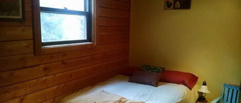 Una habitación pequeña y acogedora con una cama individual, una mesita de noche, una lámpara y una ventana con vistas a árboles.