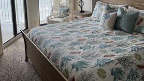 A bedroom with a large bed, a ceiling fan, a view of the ocean, and a chair by the window.