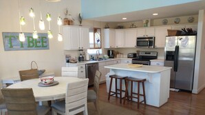 A modern kitchen with white cabinets, a central island, and a dining area with wooden chairs. - Peaceful House on South Island with Bikes- discounts on weekly summer stays! (Tybee Island)
