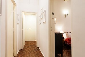 Un pasillo con suelo de madera, paredes blancas y una cama con colcha roja de lunares al fondo.