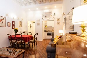 A dining area with a red tablecloth, wooden chairs, and a glass table. There is a kitchen area with white cabinets and a stove.