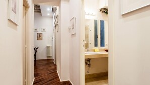 Een smalle gang met houten vloer, witte muren en ingelijste foto's. Een badkamer met wastafel, spiegel en een geel zeepdispenser is zichtbaar.