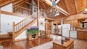 A spacious living area with a wooden staircase, a dining table, and a kitchen area. - Quiet Cabin Nestled in trees with King -hot tub, pool, pool table, fishing pond (Sevierville)
