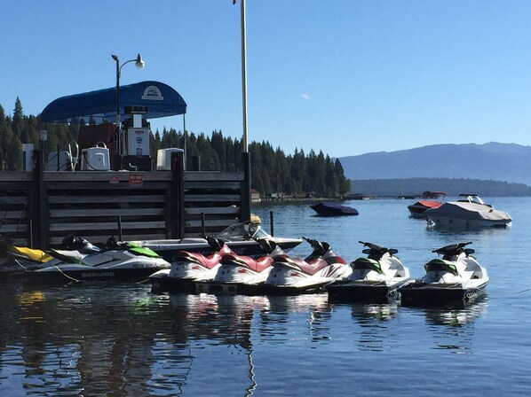 A marina with several watercraft docked, a covered area with equipment, and a forested shoreline in the background.