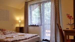 A bedroom with a bed, a nightstand, a lamp, a window with curtains, a chair, and a potted plant.