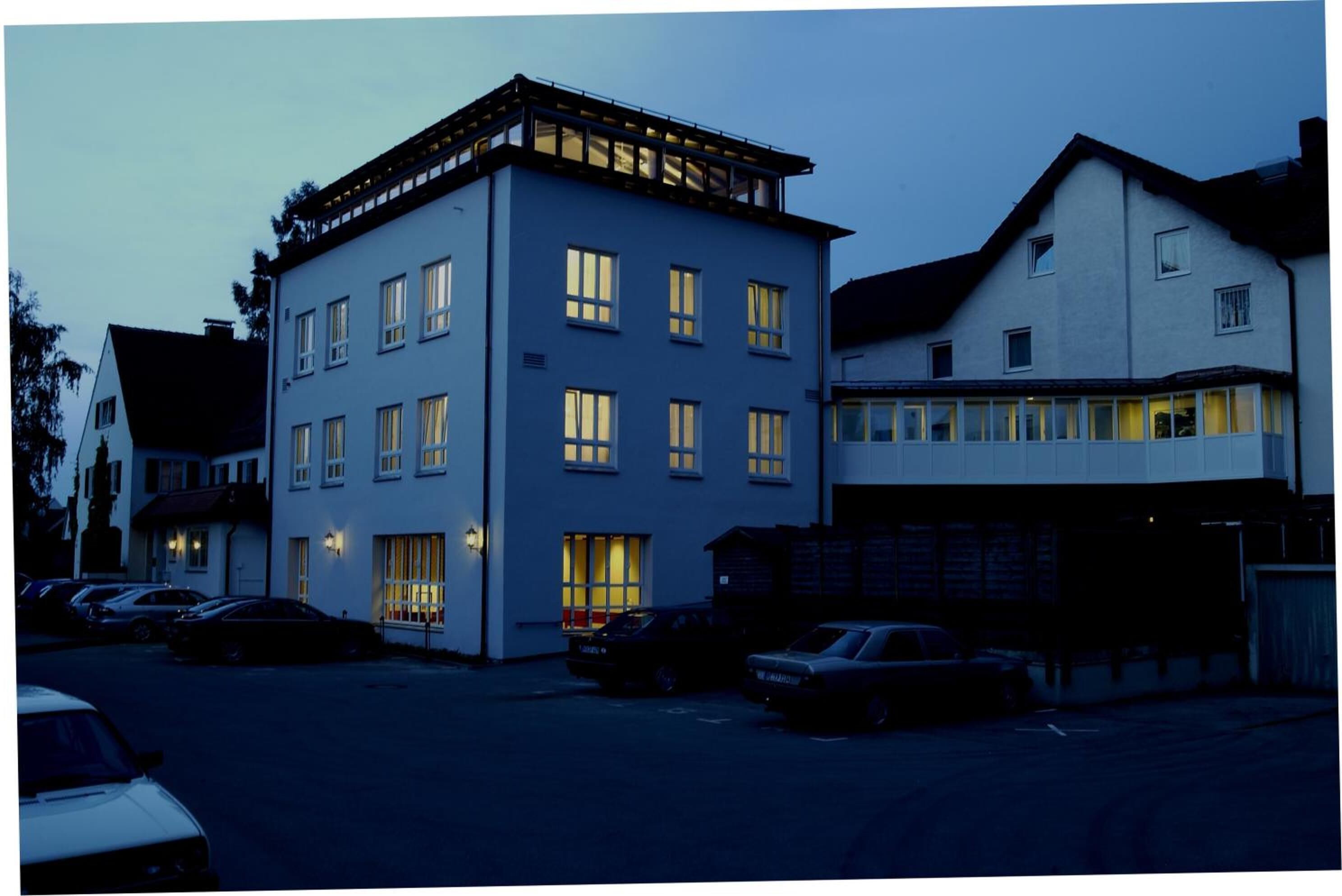 A multi-story building with several windows, a flat roof, and a parking area with cars.