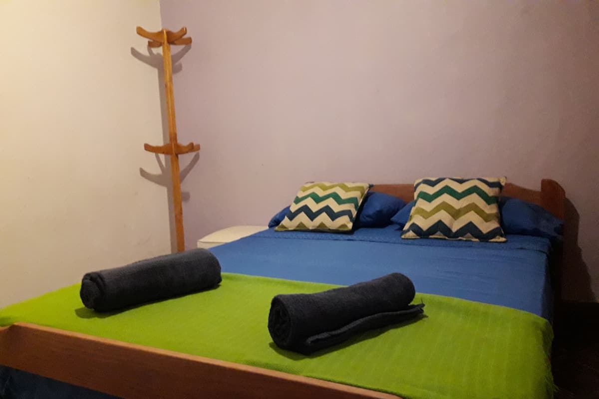 Una cama con una colcha verde y azul, dos almohadas con estampados en zigzag y dos toallas enrolladas.