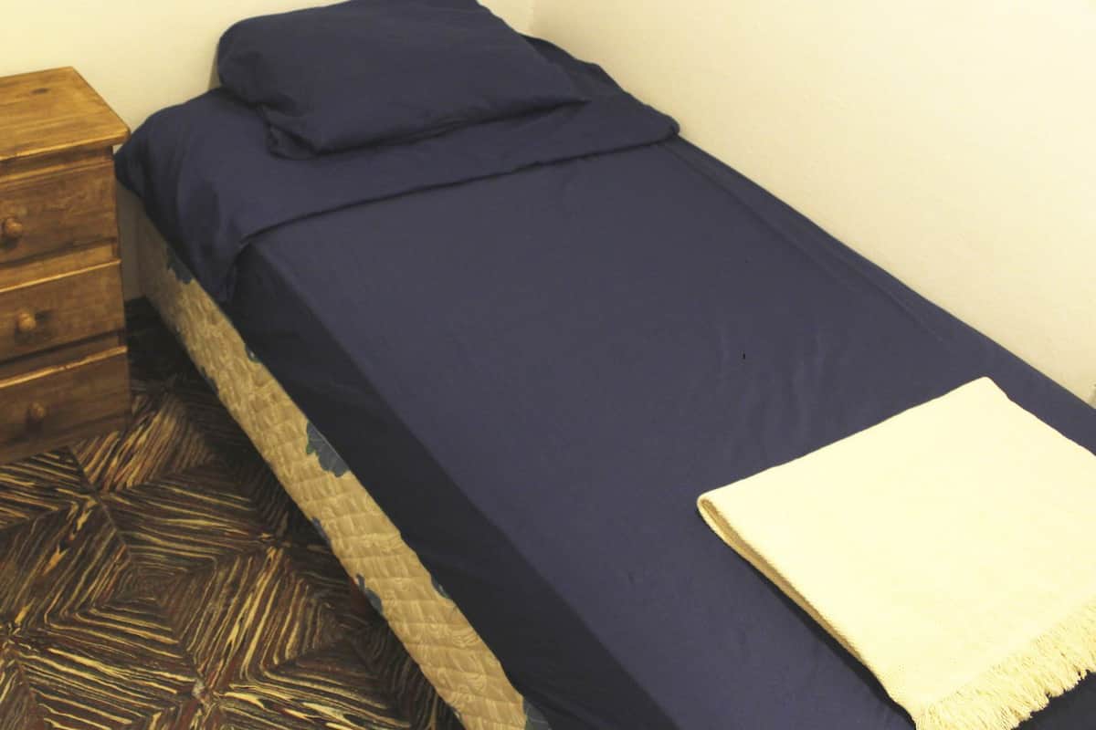 Una habitación pequeña con cama individual, un mesita de noche de madera, piso estampado y pared lisa.