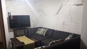 Un salon avec un canapé de couleur sombre, un téléviseur à écran plat posé sur un meuble, une petite table en bois et un coussin décoratif à motifs.