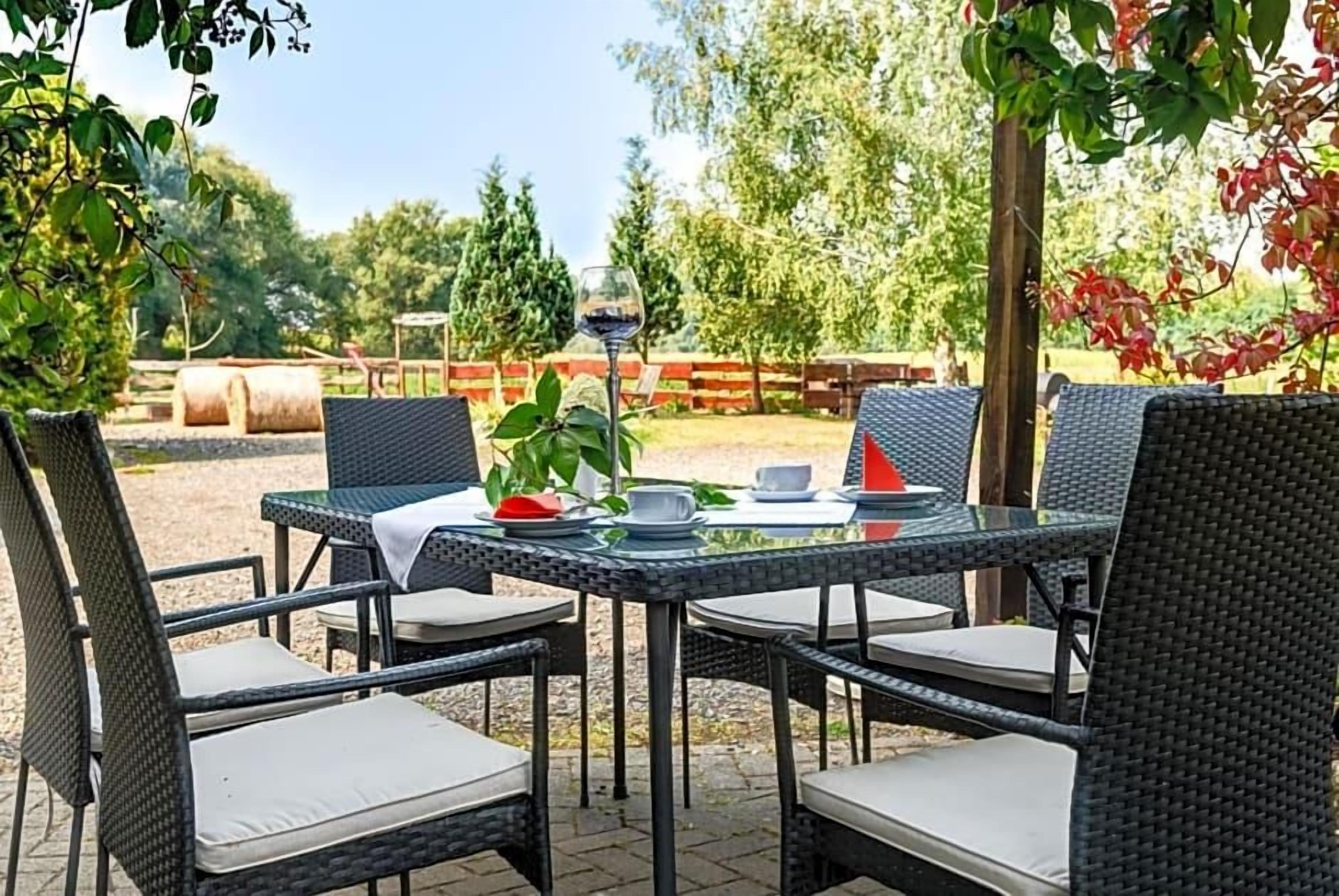 An outdoor dining area with wicker chairs and a table set for two, surrounded by greenery and a wooden fence.