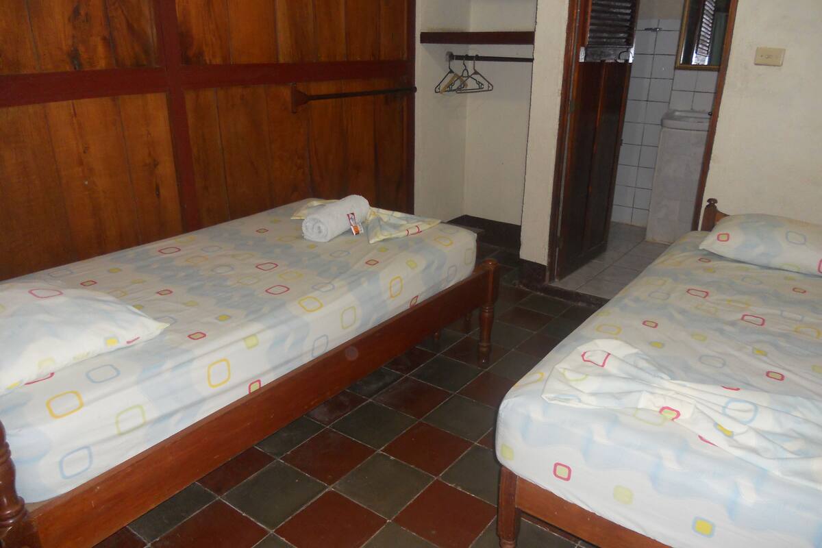 Ein Zimmer mit zwei Holzbetten, jeweils mit gemusterten Bettdecken und Kissen. Es gibt einen Holzschrank und einen Fliesenboden.