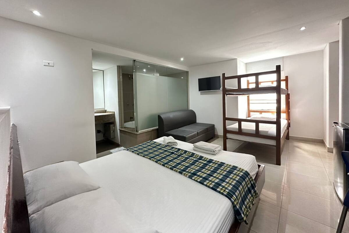 Habitación de hotel con cama, sofá, televisor, literas y zona de ducha.