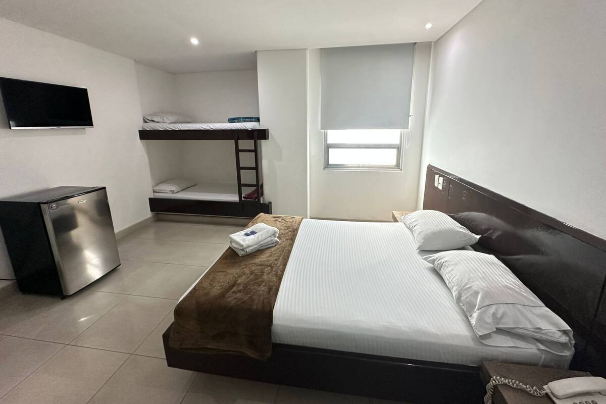 Una cama individual con cabecera marrón, una mesita de noche con un teléfono, un escritorio pequeño con una computadora, una cama litera, una ventana con persianas y un televisor montado en la pared.