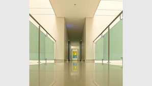A modern, well-lit hallway with glass walls and a reflective floor, leading to a brightly lit area with a yellow and blue sign. - Hotel via 45 Centro (Medellín)