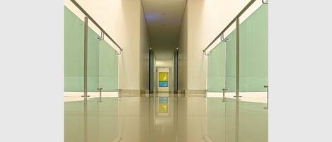 A modern, well-lit hallway with glass walls and a reflective floor, leading to a brightly lit area with a yellow and blue sign.