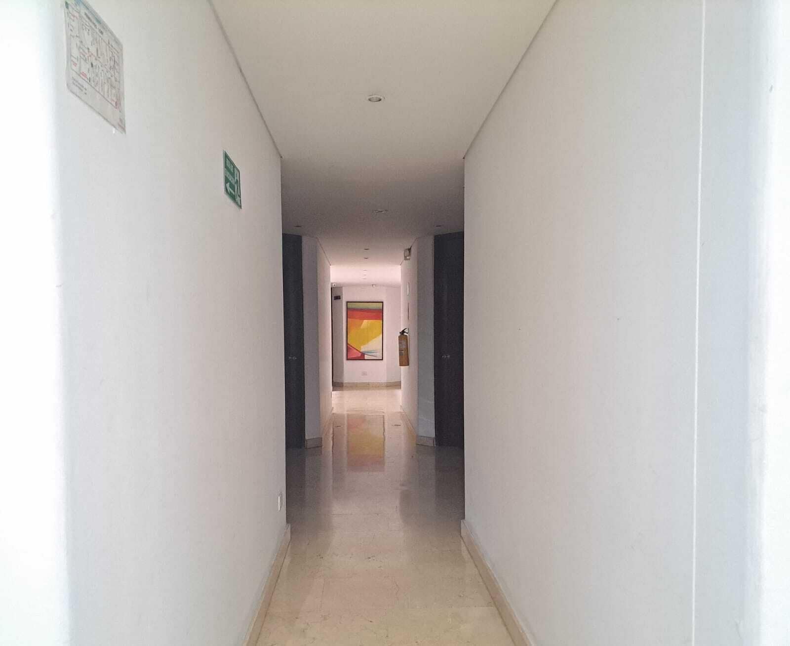Un pasillo estrecho y bien iluminado, con suelo de baldosas, un extintor de incendios montado en la pared y una puerta al final que da a otra habitación.