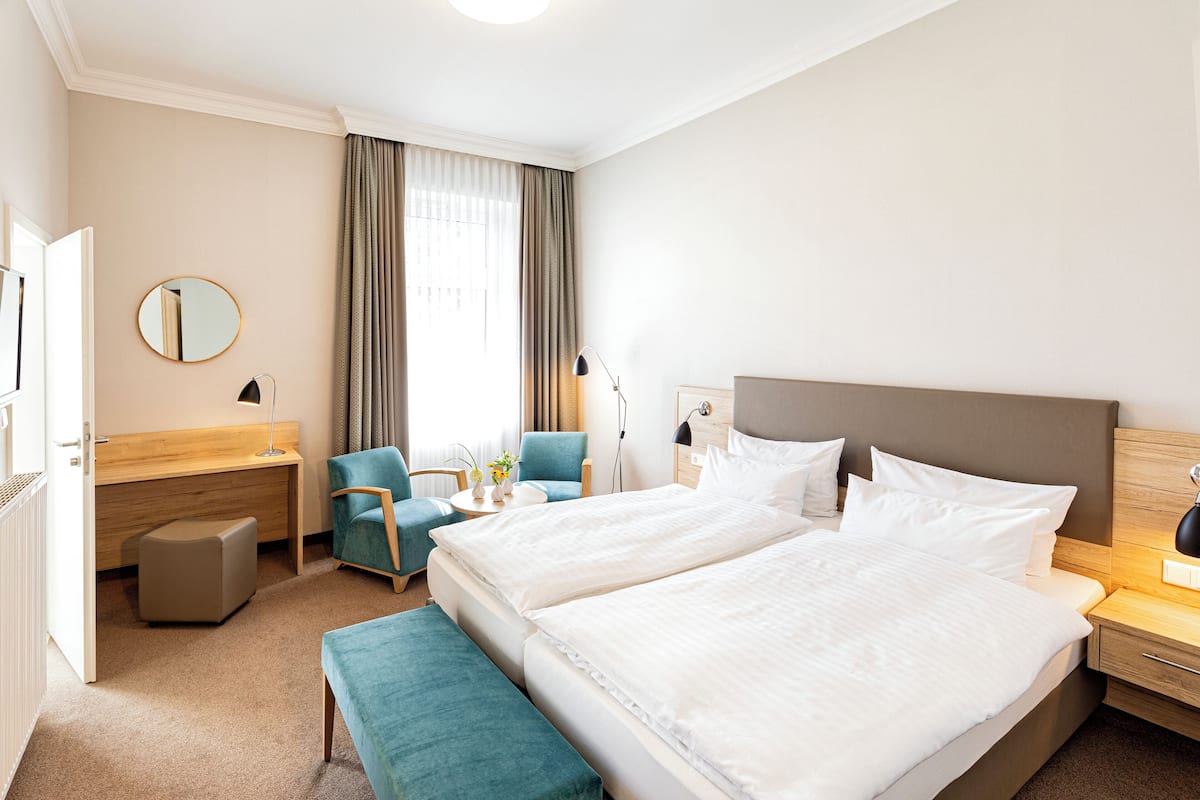 A hotel room with a large bed, a desk, a chair, a small table with flowers, and a window with curtains.
