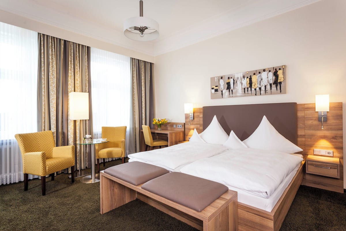 A hotel room with a large bed, two bedside tables, a desk with a chair, a small table with a lamp, and a large window with curtains.