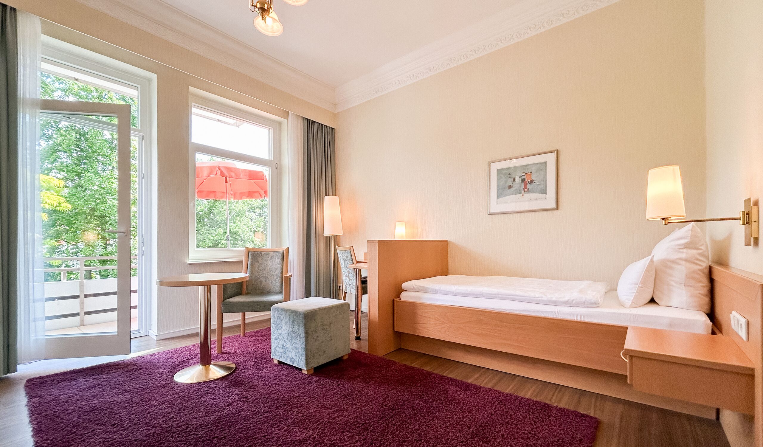 A hotel room with a bed, a small table, a chair, a window with curtains, and a view of greenery outside.