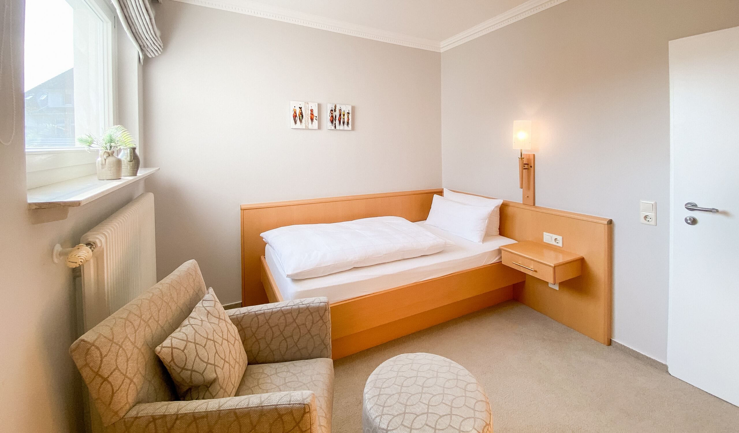Une chambre d’hôtel avec un lit simple, une table de chevet, une chaise, un petit pouf rond et une fenêtre donnant sur la rue.