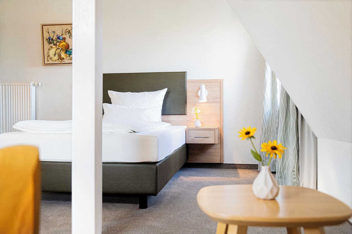 A modern hotel room with a neatly made bed, a bedside table with a lamp, and a small table with a vase of flowers.