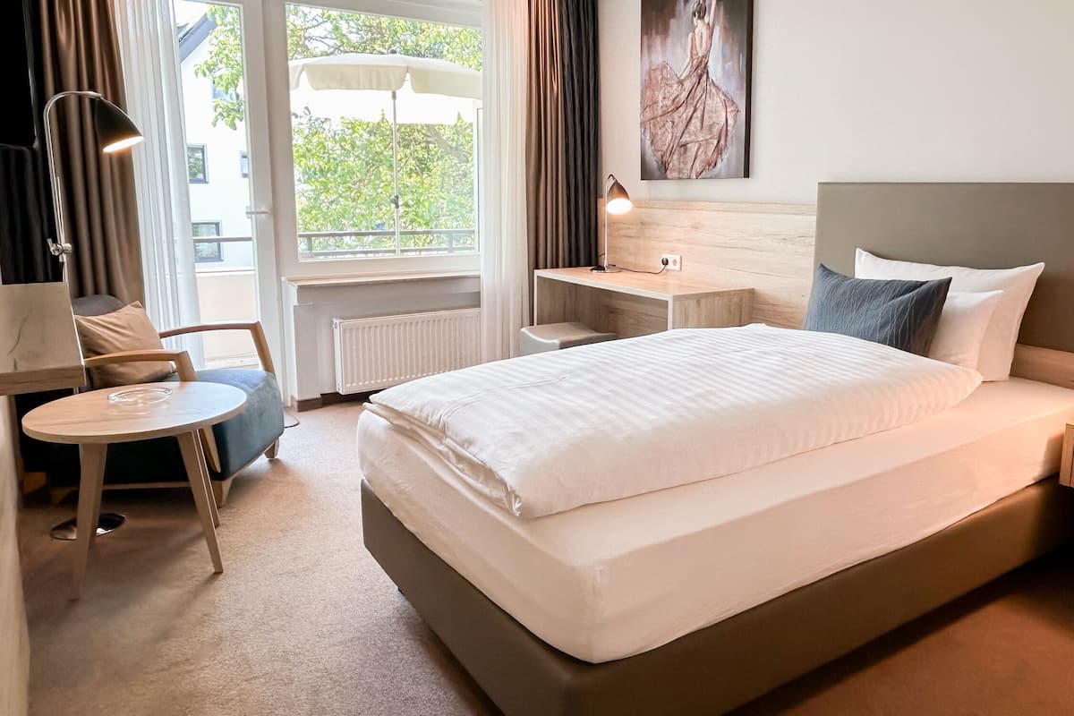 A hotel room with a large bed, a desk, a chair, and a view of greenery outside.