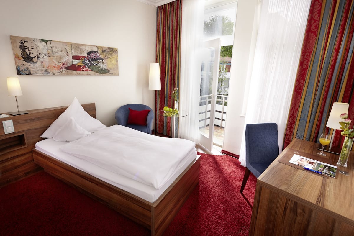 A hotel room with a bed, a desk, a chair, a lamp, a painting, and a view of the outdoors.