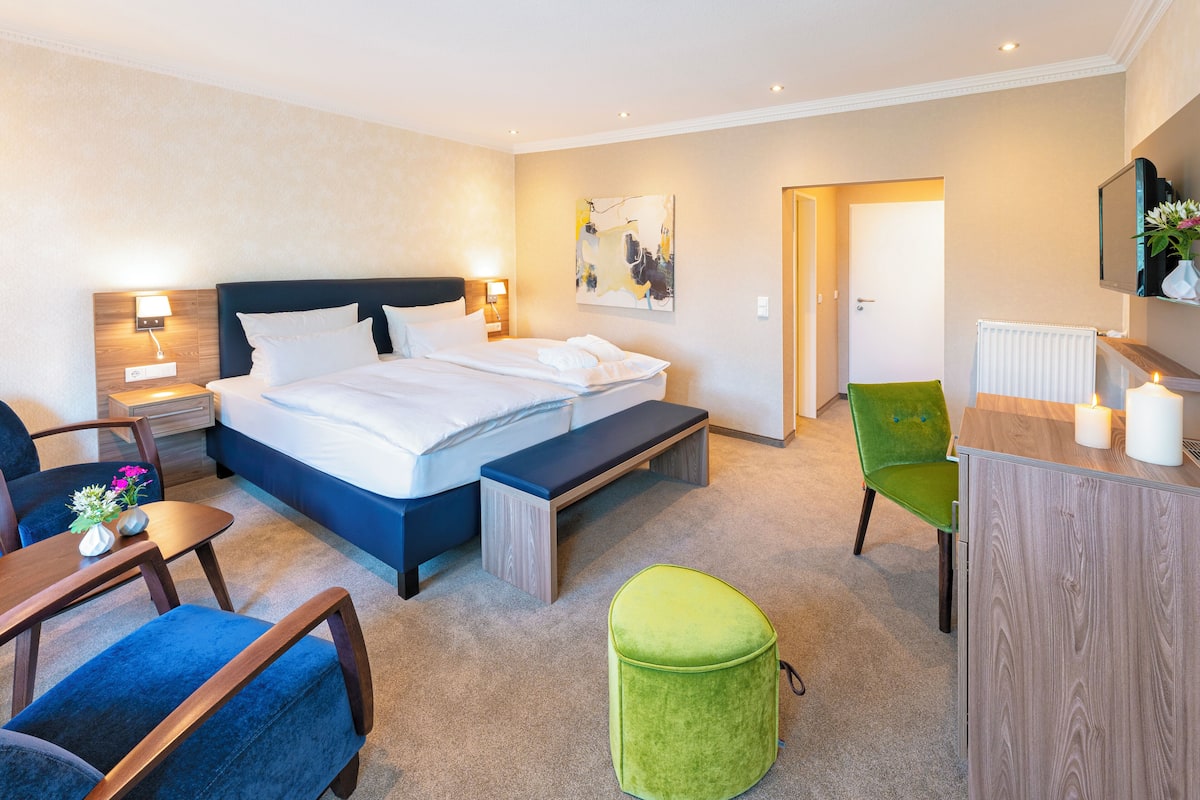 A modern hotel room with a large bed, bedside tables, a desk, a green ottoman, and a TV on a wooden cabinet.