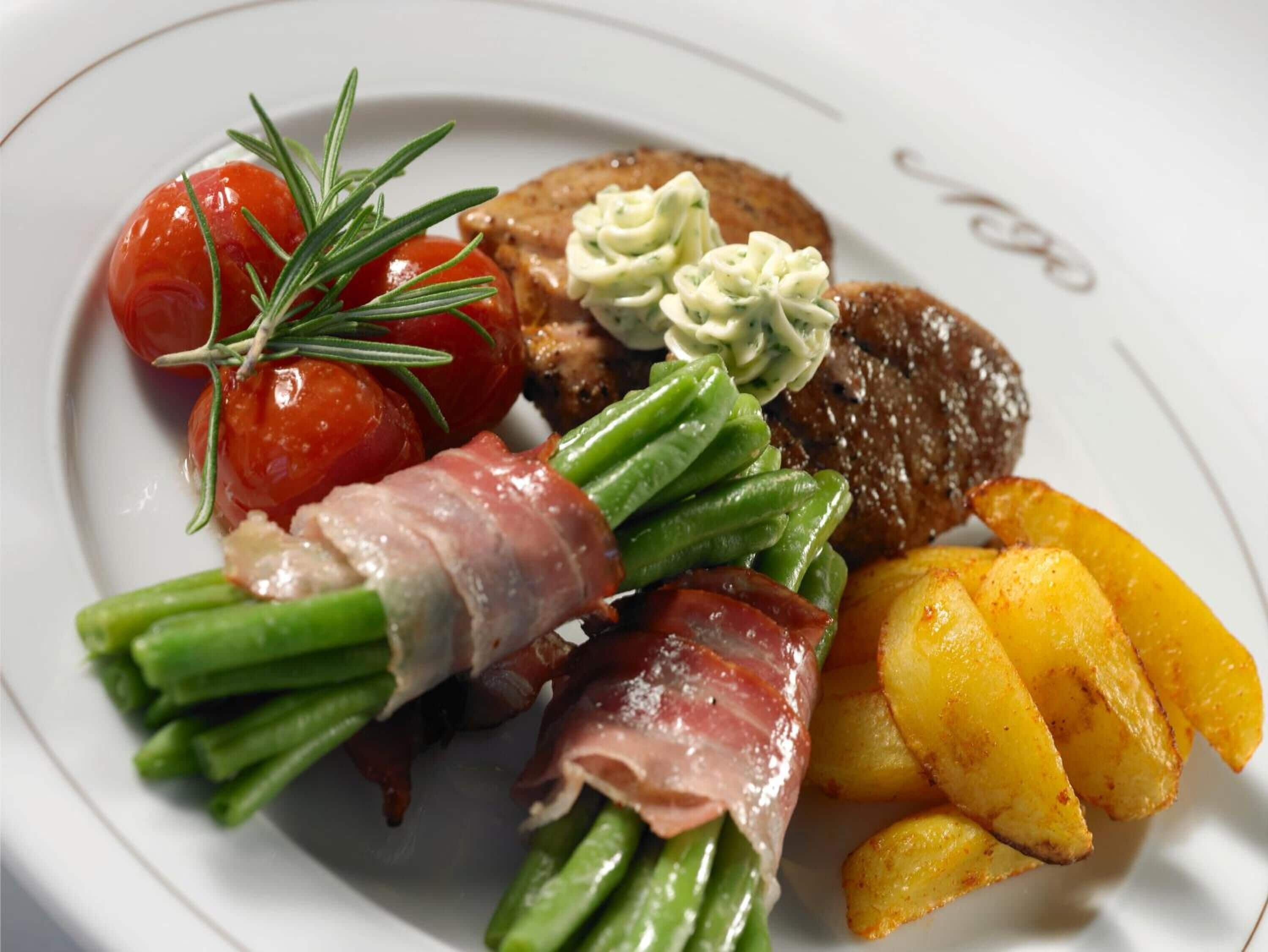 Een bord met diverse gerechten, waaronder een steak, groene bonen, tomaten en geroosterde aardappelen.