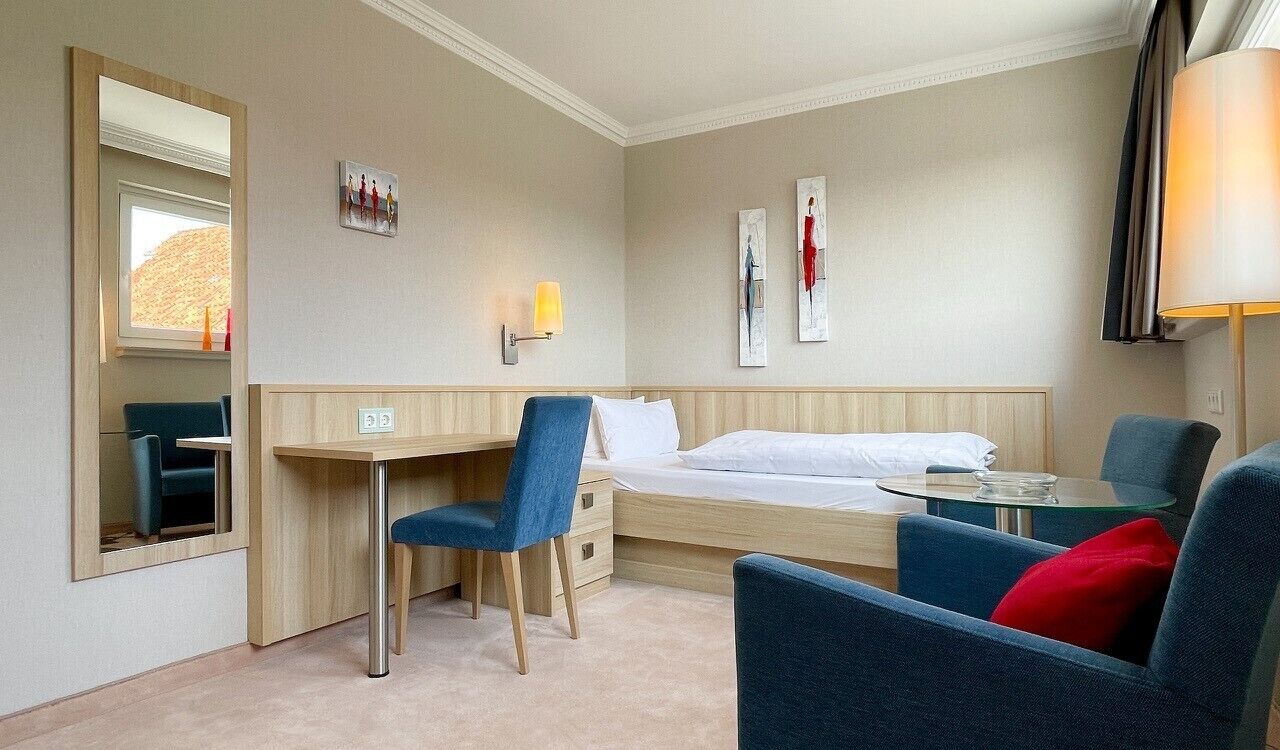 Ein Hotelzimmer mit einem Bett, einem Schreibtisch, zwei Stühlen, einem kleinen Tisch, einer Lampe und Kunstwerken an den Wänden.