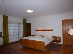 A bedroom with a wooden bed, a bedside table, a nightstand, a lamp, a window with curtains, and a door.