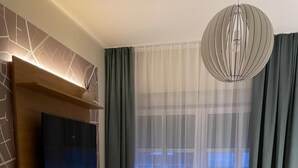 A modern bedroom with a flat-screen TV mounted on a wall, a grey upholstered chair, a large bed with white bedding, and a large window with sheer curtains. - Haselhoff (Coesfeld)