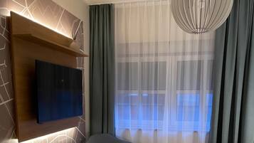 Ein modernes Schlafzimmer mit einem an der Wand befestigten Flachbildfernseher, einem grau gepolsterten Sessel, einem großen Bett mit weißer Bettwäsche und einem großen Fenster mit transparenten Vorhängen.