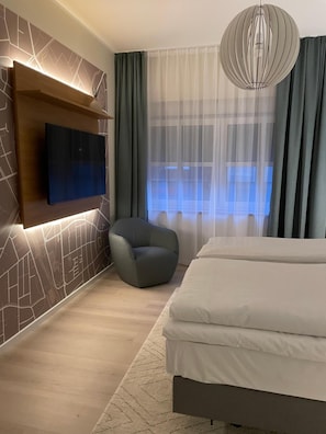A modern bedroom with a flat-screen TV mounted on a wall, a grey upholstered chair, a large bed with white bedding, and a large window with sheer curtains. - Haselhoff (Coesfeld)