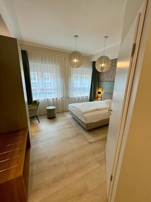 Une chambre à coucher moderne, avec un lit, deux lampes suspendues, un fauteuil et une vue sur la rue grâce à de grandes fenêtres.