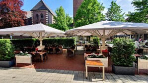 An outdoor café with tables and chairs under umbrellas, surrounded by greenery and a building with a tower. - Haselhoff (Coesfeld)