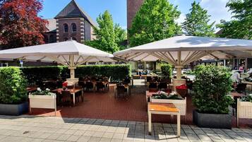 Ein Straßencafé mit Tischen und Stühlen unter Sonnenschirmen, umgeben von Grünpflanzen und einem Gebäude mit einem Turm.