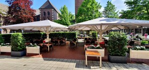 An outdoor café with tables and chairs under umbrellas, surrounded by greenery and a building with a tower. - Haselhoff (Coesfeld)