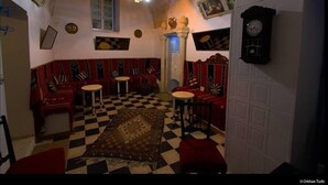 Een kamer met een geruite vloer, rode en zwarte gestreepte banken, een kroonluchter en een klok aan de muur.